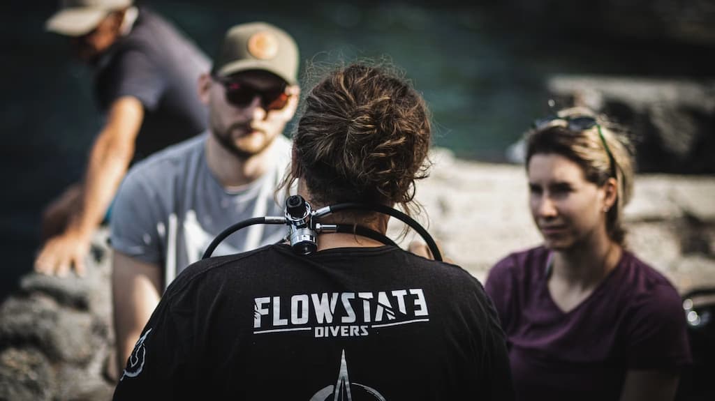 Three divers in FlowState Divers shirts talking on a rocky shore after a dive.